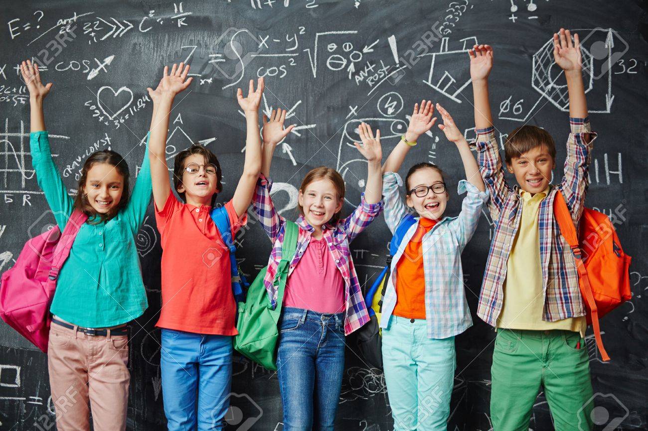 happy classmates raising hands while standing against blackboard