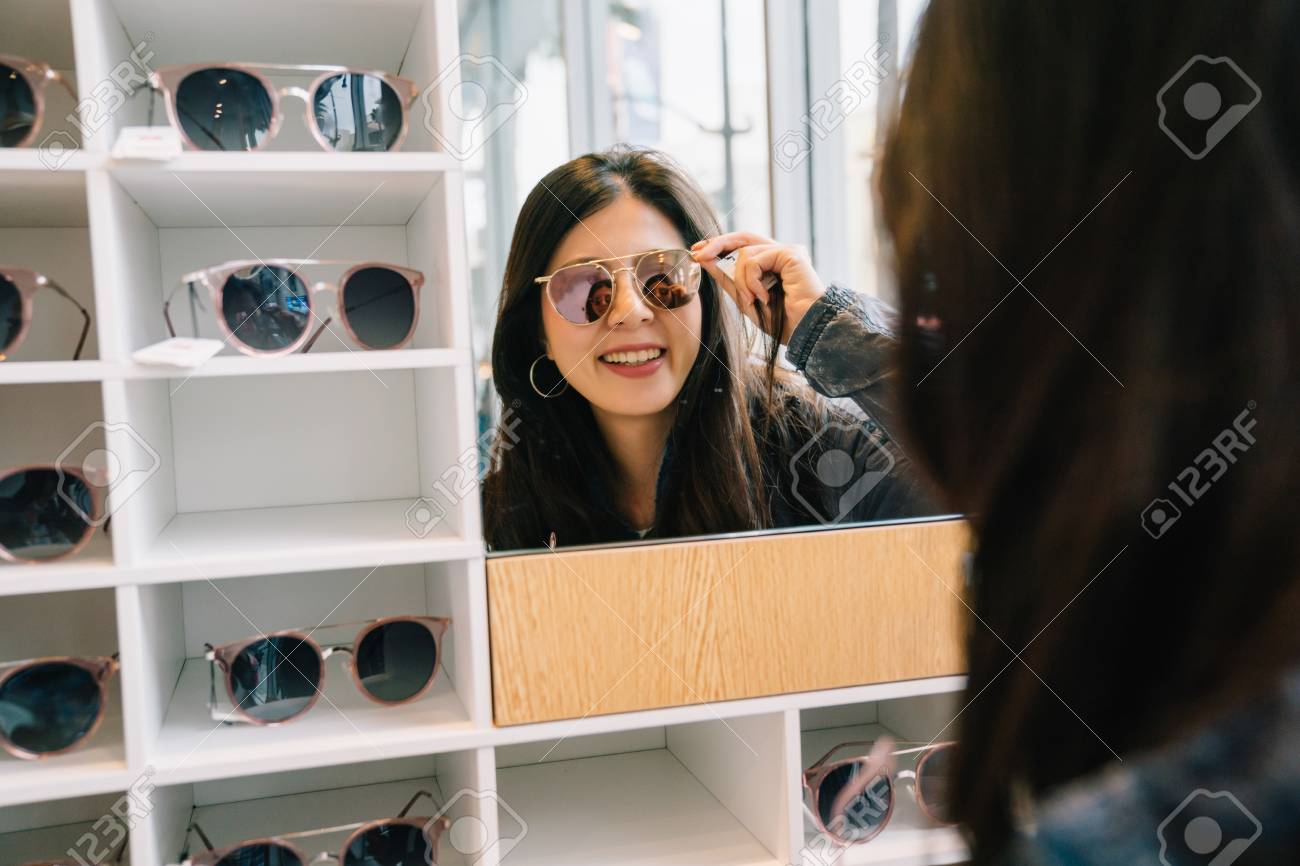 attractive woman trying sunglasses in the glasses shop and looking in the mirror. Asian lady shopping lifestyle. beautiful girl in suitable sunglasses. - 109943562 attractive woman trying sunglasses in the glasses shop and looking in the mirror. Asian lady shopping lifestyle. beautiful girl in suitable sunglasses. - 109943562
