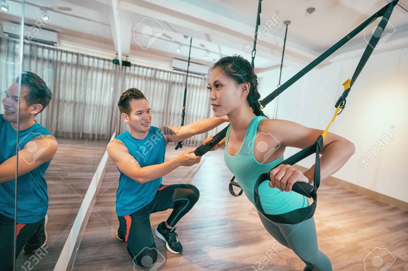 개인 코치와 함께 체육관에서 TRX 훈련을 하는 젊은 아시아 여성. 탄력 있는 밧줄로 스트레칭 운동을 하는 스포티한 소녀. 횡단 밴드를  사용하여 슬링 훈련에서 트레이너와 함께 스포츠 레이디. 로열티 무료 사진, 그림, 이미지