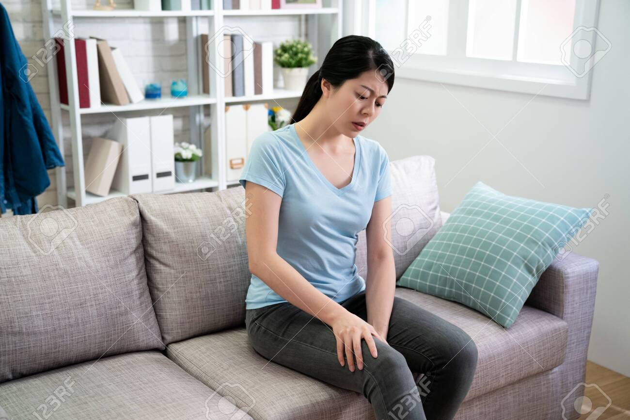 Closeup young woman sitting on sofa and feeling knee pain and massage her knee at home. Healthcare and medical concept. asian girl housewife sitting on couch frowning looking at hurting legs - 121240486 Closeup young woman sitting on sofa and feeling knee pain and massage her knee at home. Healthcare and medical concept. asian girl housewife sitting on couch frowning looking at hurting legs - 121240486