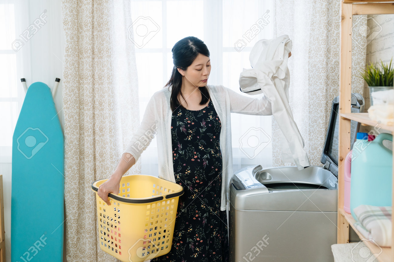 人妻の洗濯物 アジアの中国人妊婦が洗濯機に服を追加し、明るい室内で洗濯かごを持っています。中に夫のシャツを入れている若い産婦人妻。家事をしている魅力的な未来のお母さん。の写真素材・画像素材  Image 164245561