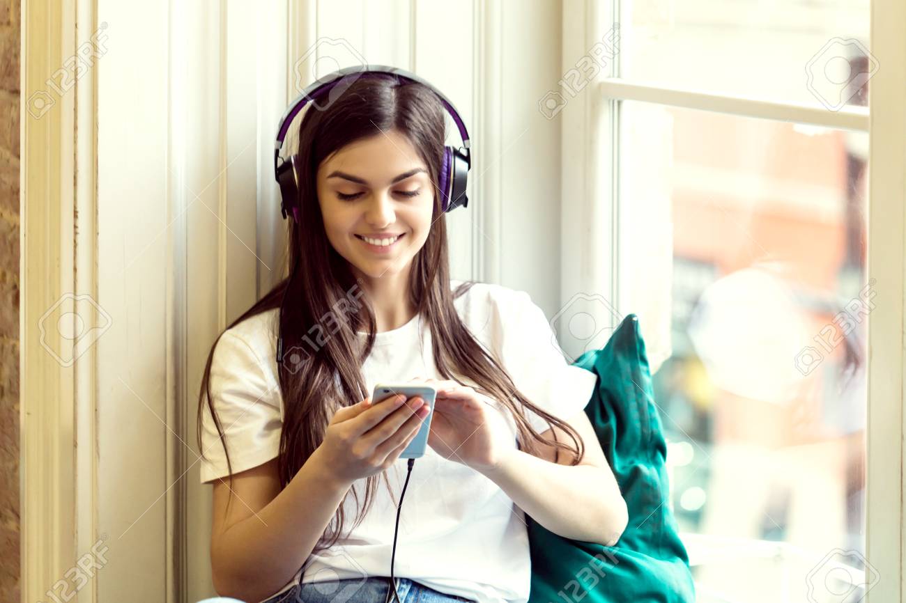 Long hair girl wears headphones and white t-shirt listen to music sitting on wide windowsill - 100125674 Long hair girl wears headphones and white t-shirt listen to music sitting on wide windowsill - 100125674
