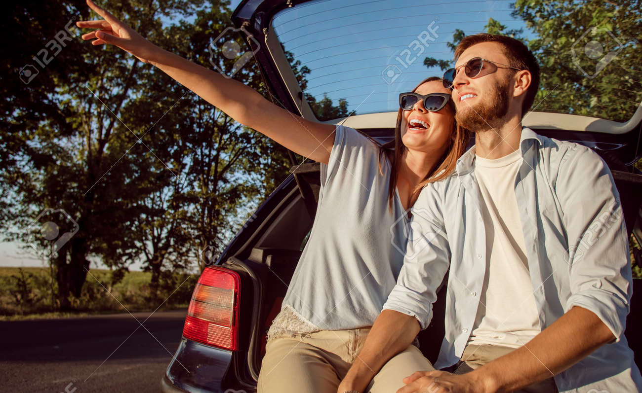 Young cheerful couple admires the countryside views while sitting in the car trunk in the rays of sunset sun - 145147751 Young cheerful couple admires the countryside views while sitting in the car trunk in the rays of sunset sun - 145147751