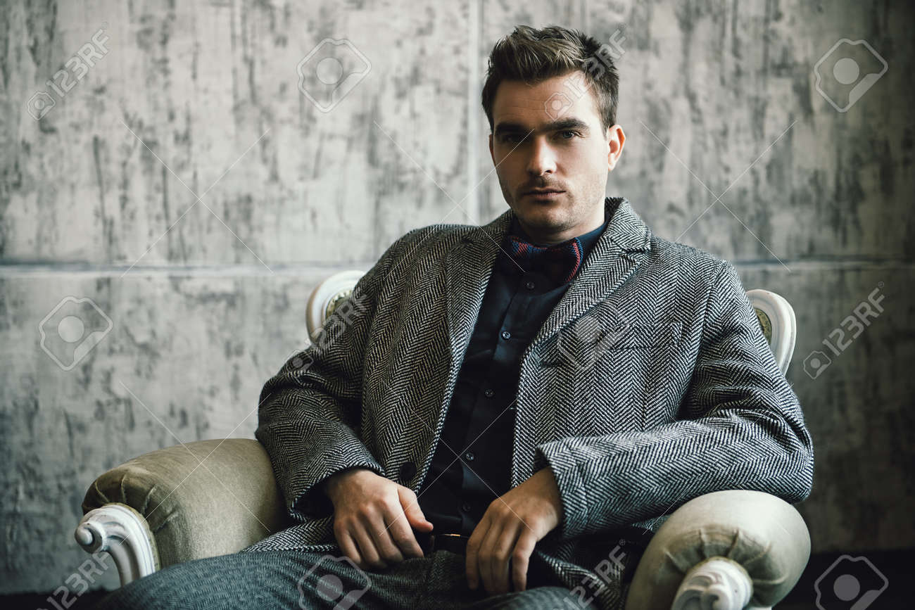 A portrait of a fashionable young man posing in the armchair over the grey background. Formal fashion for men. - 120873004 A portrait of a fashionable young man posing in the armchair over the grey background. Formal fashion for men. - 120873004