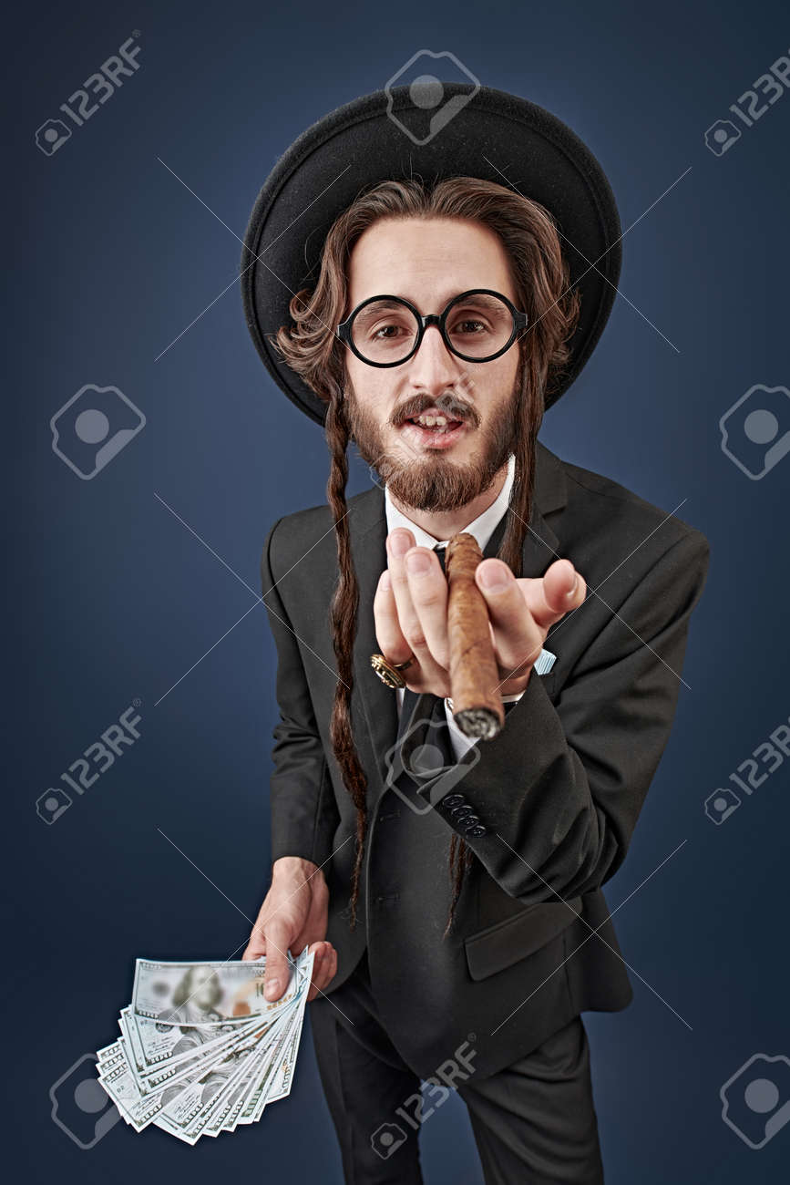 부유한 유태인 개념. 손가락에 금반지를 끼고 지폐 다발을 들고 시가를 피우는 부유한 유대인의 초상화. 어두운 파란색 배경에 촬영  스튜디오입니다. 로열티 무료 사진, 그림, 이미지 그리고 스톡포토그래피. Image 172040645