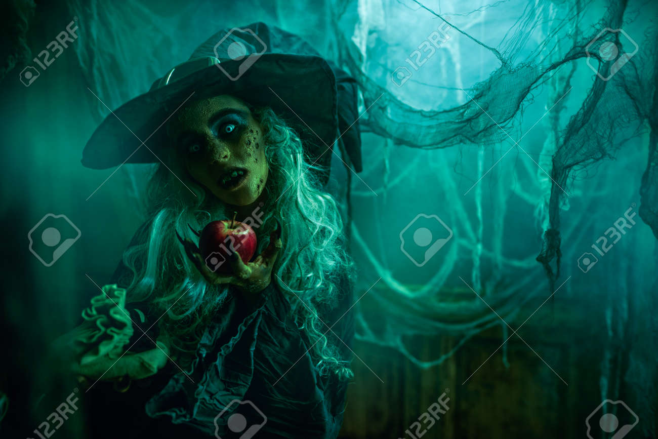 Halloween. A Terrible Wicked Witch Holds A Poisoned Apple In Her Hands,  Standing In Her Lair Among The Cobwebs. Evil Black Magic. Stock Photo,  Picture and Royalty Free Image. Image 189691888., image size:1300x867