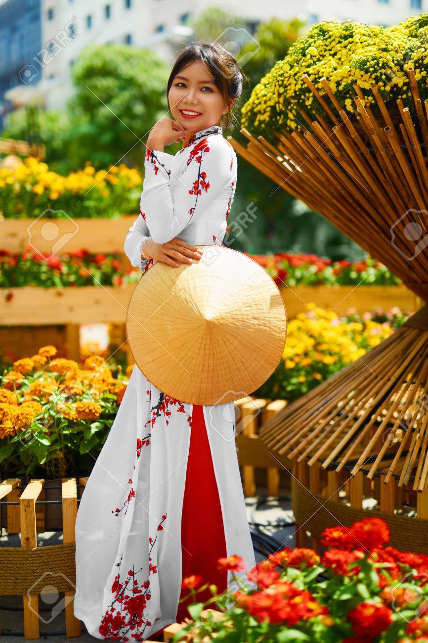 Ropa Tradicional De Vietnam, Asia. La Asiática Hermosa Mujer Vestida Con Vestido Tradicional Nacional Dai (vestuario), Vietnamita Sombrero (non La, Sombrero De La Hoja) En Jardín De Flores. Cultura. Fotos,