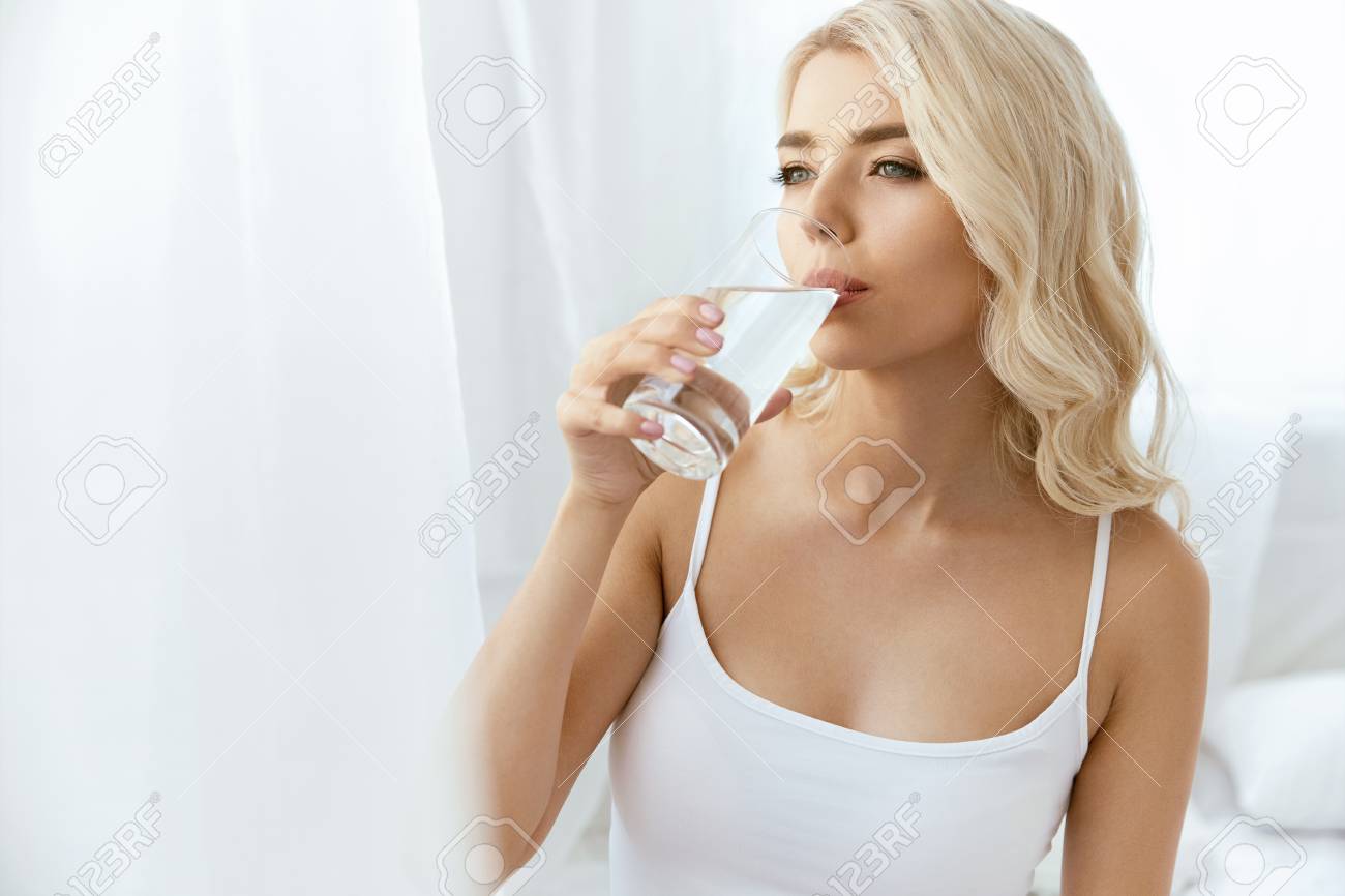 飲料水 コップ一杯の水を持つ女性 明るいインテリアできれいな水を飲む美しい幸せな女性の肖像画 健康的なダイエットコンセプト 高品質の画像 の写真素材 画像素材 Image