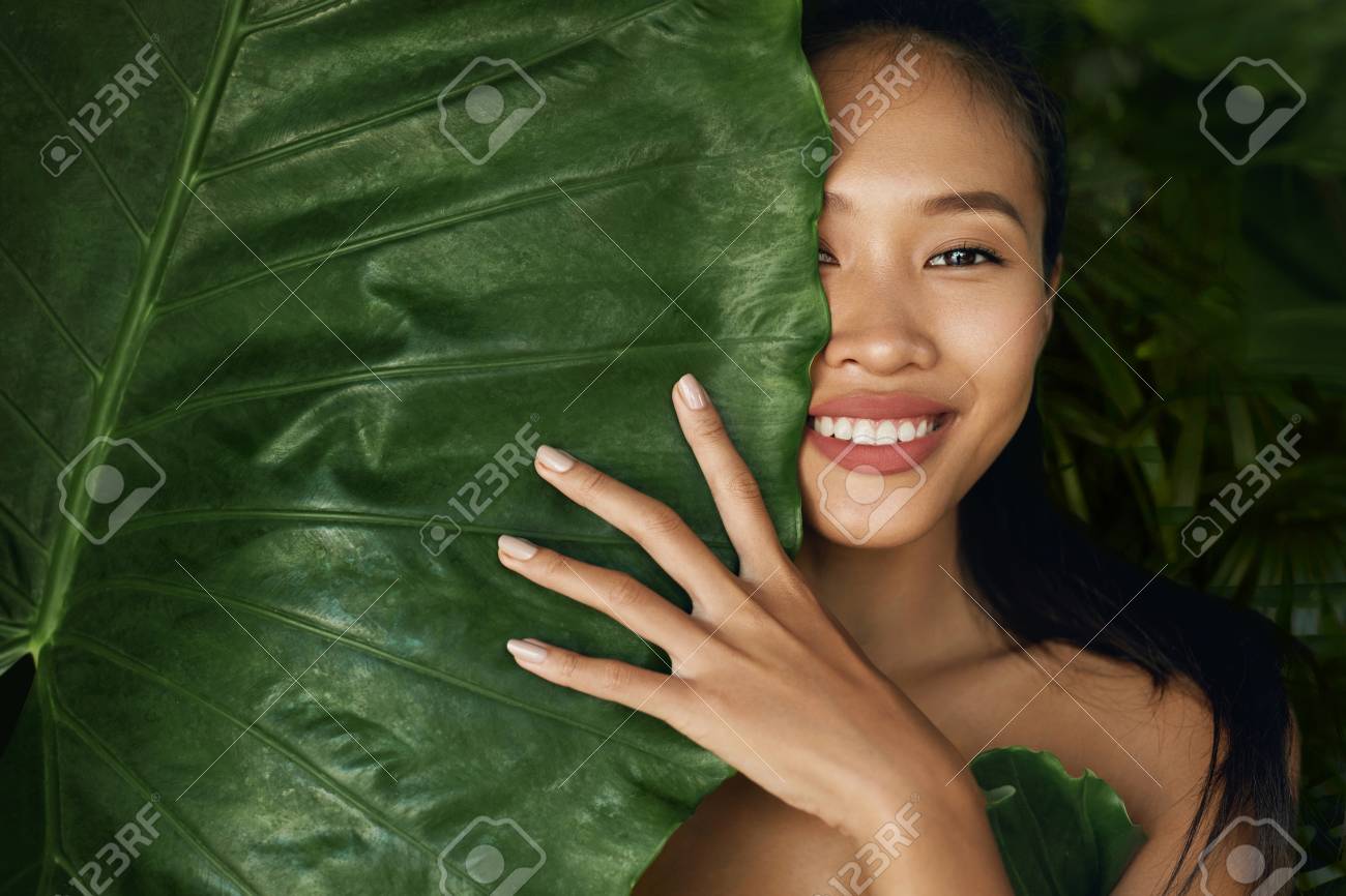 Rostro De Belleza. Modelo De Mujer Con Maquillaje Natural Y Piel Sana  Detrás De La Planta De Hojas Verdes. Retrato De Hermosa Chica Asiática Con  Uñas Desnudas, Labios Grandes Y Sonrisa Sexy