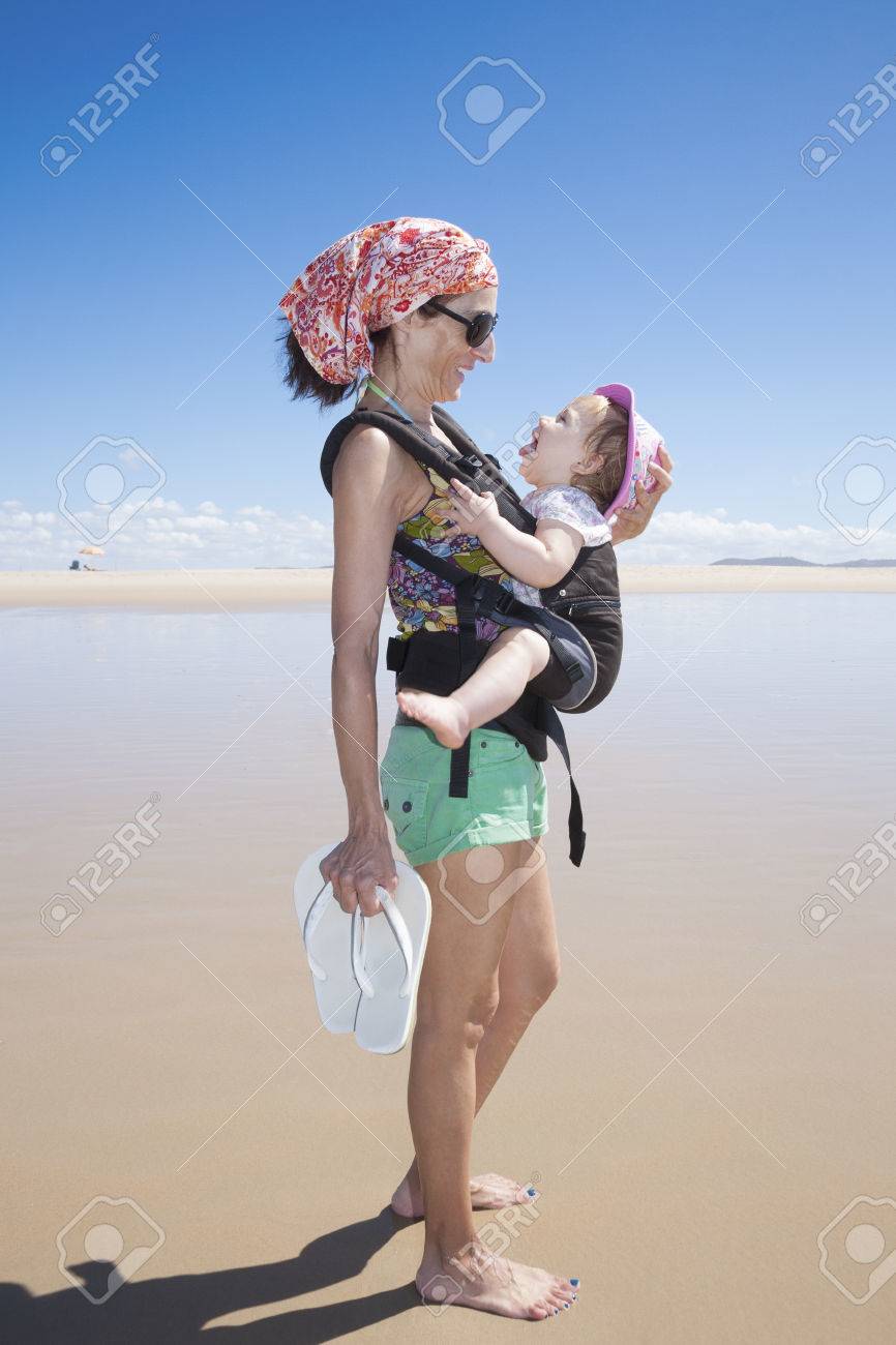 Madre Con Pañuelo Pantalones Cortos Verdes Que Bebé De Un Año Con El Sombrero De Color Rosa En La Mochila Para Caminar Frente A Una Playa Fotos, Retratos, Imágenes