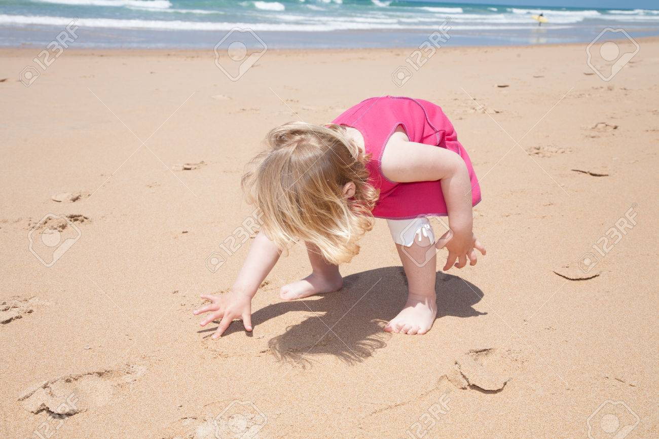 Deux Ans Fille Blonde Avec Une Robe Rose Accroupir Et écrire Avec Le Doigt Sur Plage De Sable Doré Avec La Mer Derrière