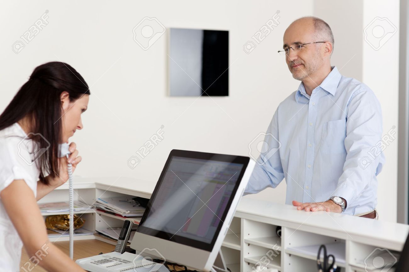 Mature male patient looking at female receptionist using landline phone and computer at reception in dentist's clinic - 21246962 Mature male patient looking at female receptionist using landline phone and computer at reception in dentist's clinic - 21246962