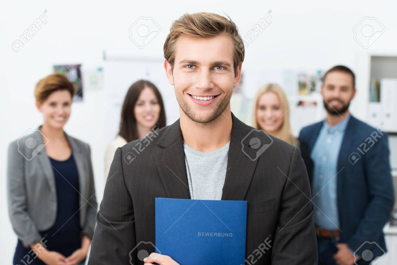成功した笑顔若い男性求職者彼の新しい仕事の同僚やビジネス チームの前でポーズ彼の履歴書と青いファイルの保持 の写真素材 画像素材 Image