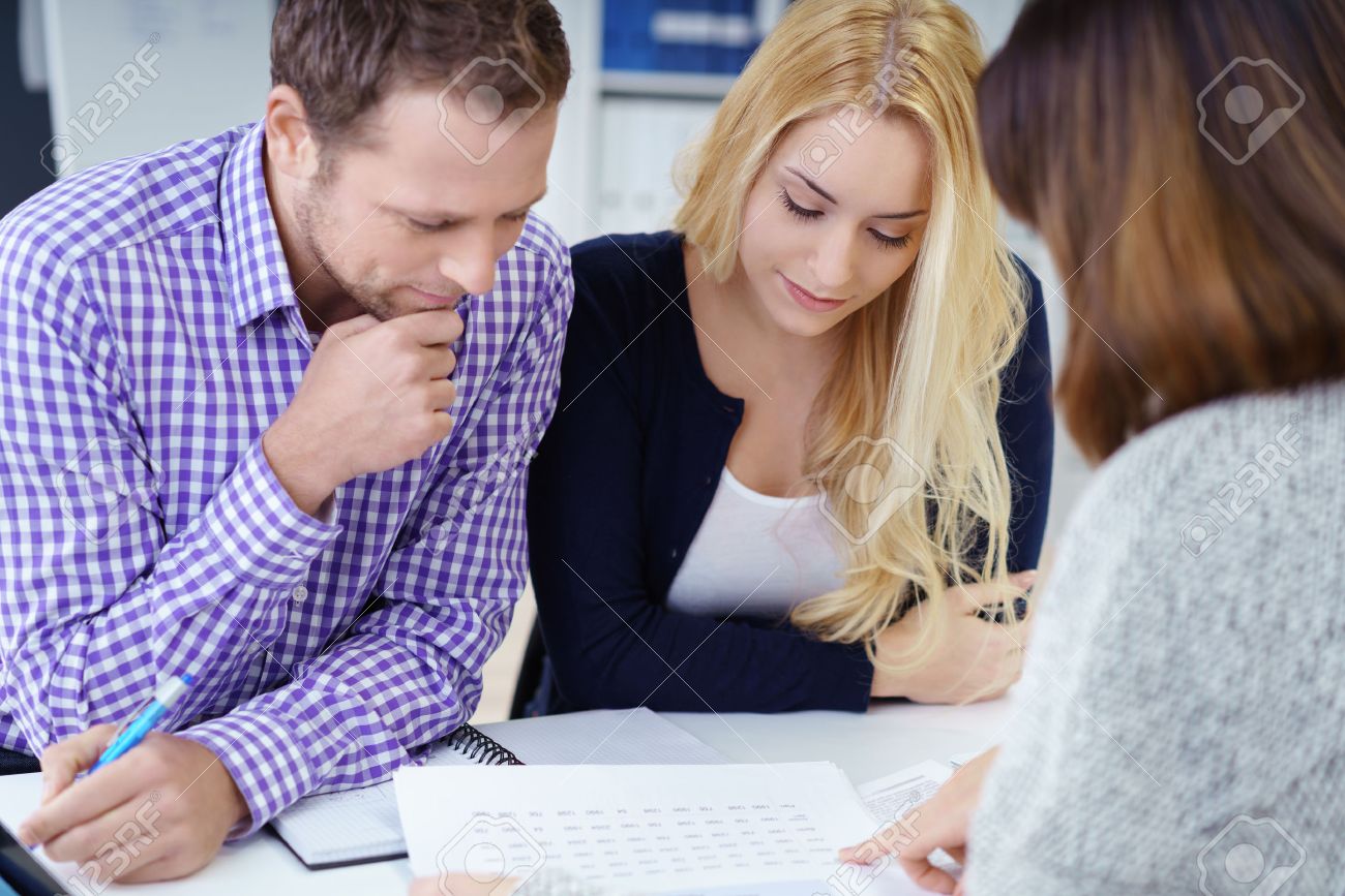 Broker giving a presentation to a young couple in her office leaning over the desk to explain paperwork to them - 51501459 Broker giving a presentation to a young couple in her office leaning over the desk to explain paperwork to them - 51501459
