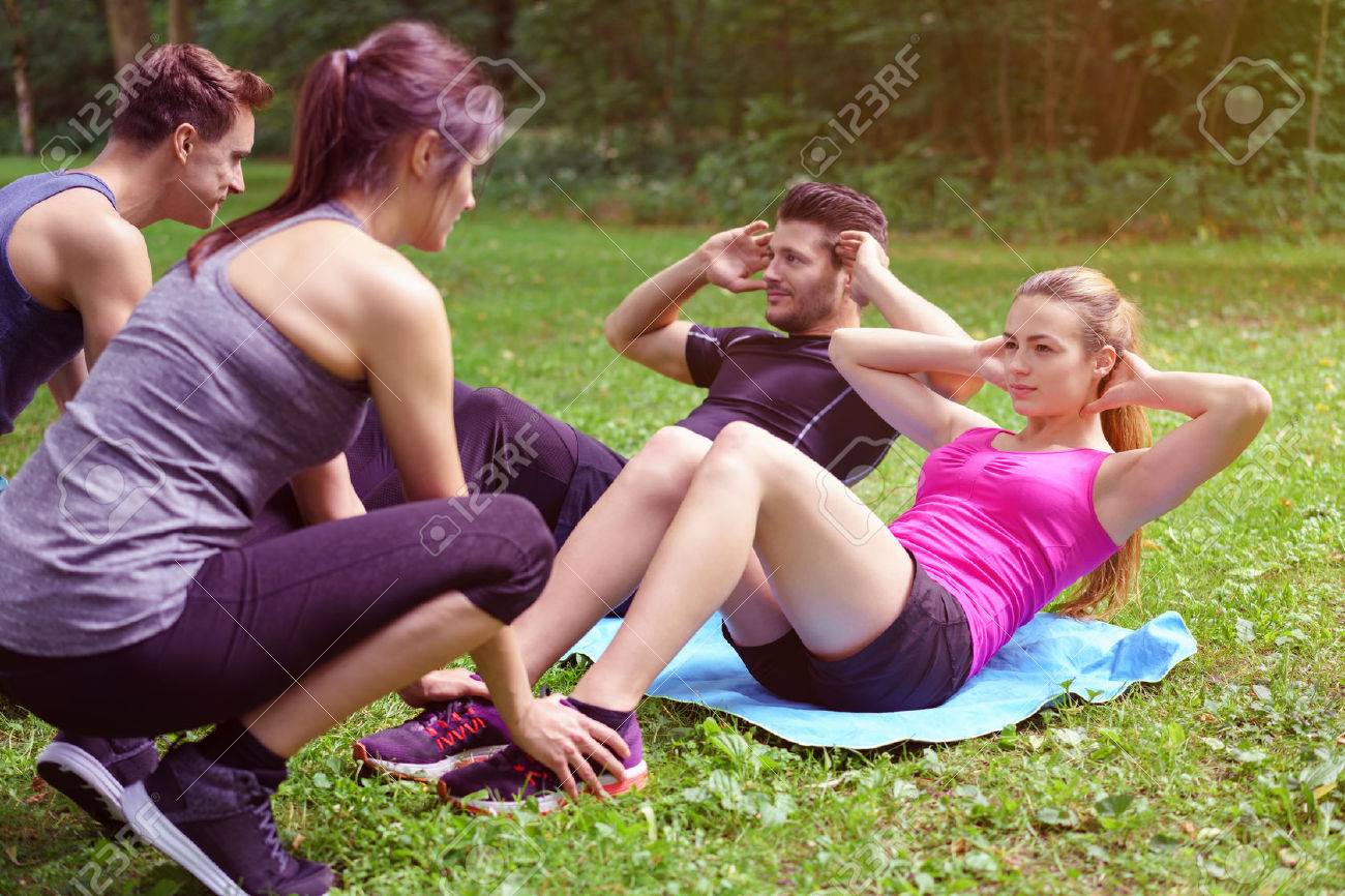 Two young couples working out in a park with one couple doing sit ups assisted by the second man and woman holding their feet - 61146892 Two young couples working out in a park with one couple doing sit ups assisted by the second man and woman holding their feet - 61146892