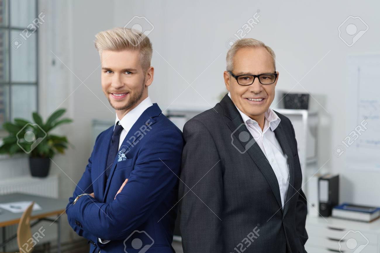 Confident successful business partners posing together with a stylish young man and his older mentor standing back to back smiling at the camera - 68433247 Confident successful business partners posing together with a stylish young man and his older mentor standing back to back smiling at the camera - 68433247