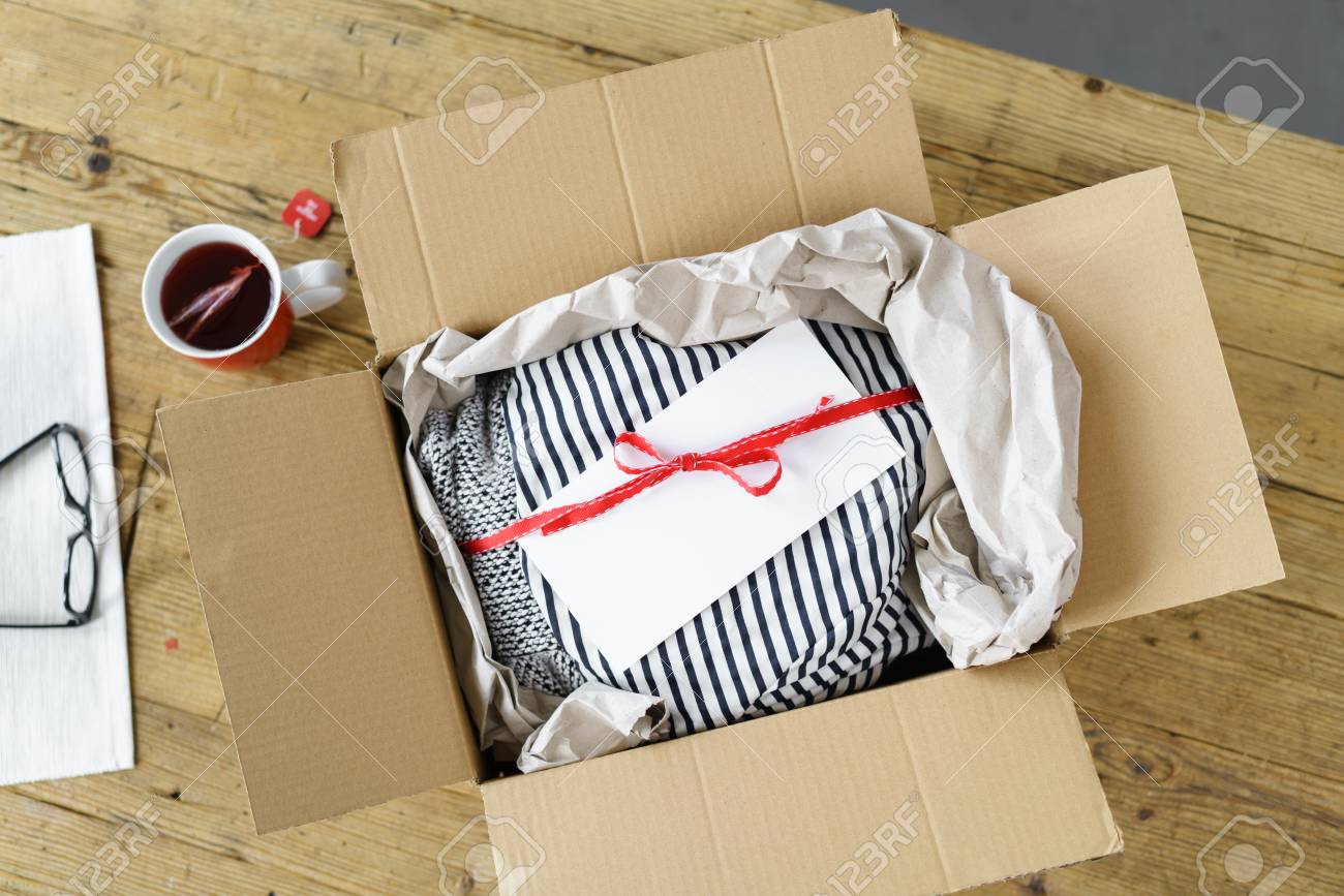 Abrió Caja De Cartón Con Un Regalo Ropa De Boutique De Moda Atados Con Una Cinta Roja Con Un Blanco Sobre Blanco, Vista Desde Arriba Sobre Una Mesa De Madera Con