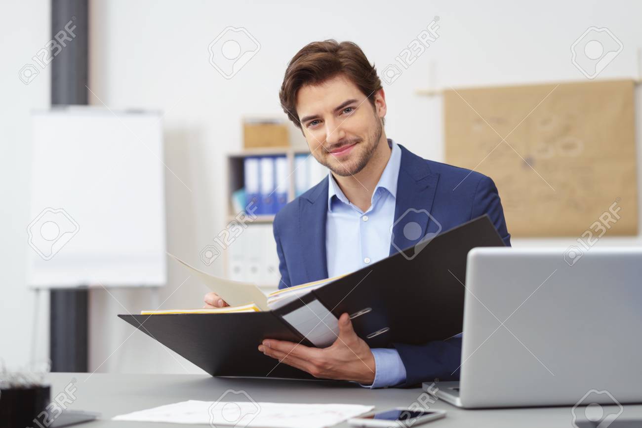Handsome businessman with a friendly smile looking at the camera as he sits holder on open office binder with paperwork - 75183562 Handsome businessman with a friendly smile looking at the camera as he sits holder on open office binder with paperwork - 75183562