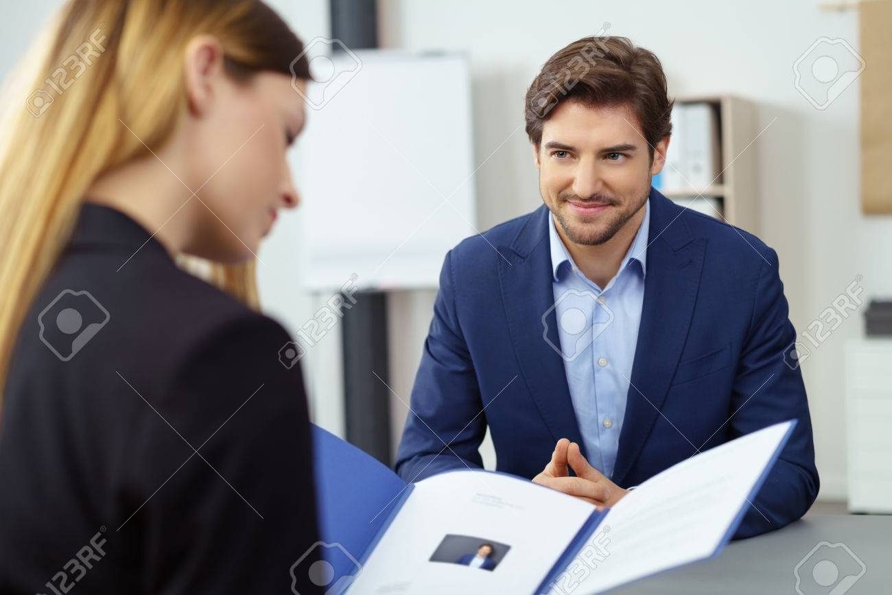 Handsome young businessman in a job interview smiling as he watches a female business executive reading his CV in a file - 76674016 Handsome young businessman in a job interview smiling as he watches a female business executive reading his CV in a file - 76674016