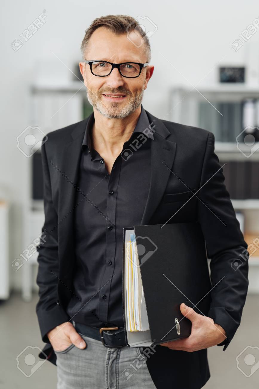 Successful businessman carrying an office binder under his arm as he stands smiling at the camera with a thoughtful expression - 94779348 Successful businessman carrying an office binder under his arm as he stands smiling at the camera with a thoughtful expression - 94779348
