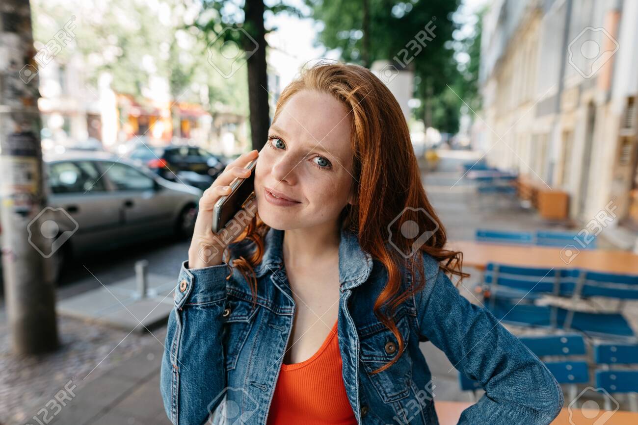 Thoughtful young woman listening to a call on her mobile phone in a quiet street in town in close up smiling quietly at the camera - 154410525 Thoughtful young woman listening to a call on her mobile phone in a quiet street in town in close up smiling quietly at the camera - 154410525