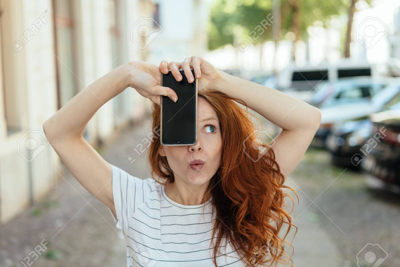 Young woman pulling a quirky face pursing her lips and looking aside as she holds up her blank mobile phone in town - 154410533 Young woman pulling a quirky face pursing her lips and looking aside as she holds up her blank mobile phone in town - 154410533