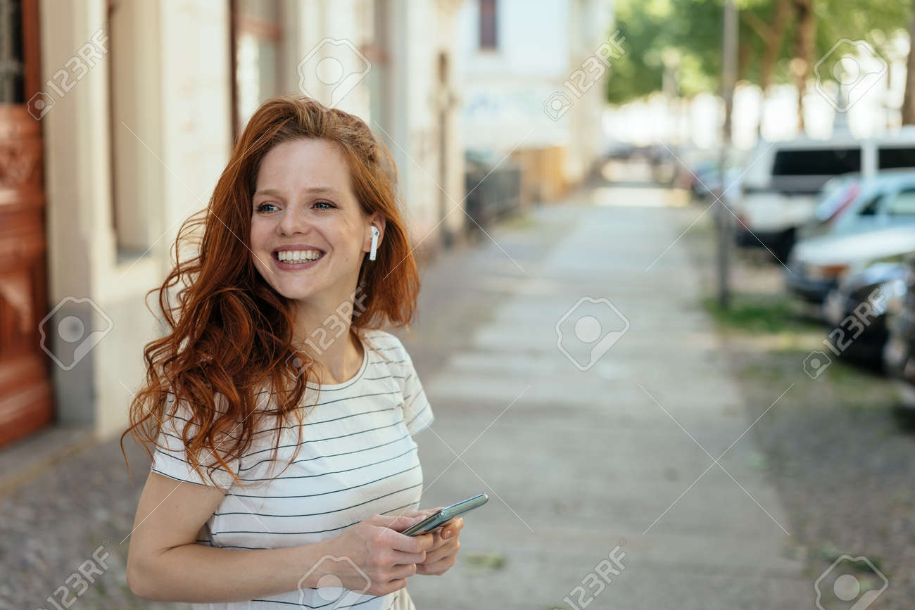 Happy young woman turning aside with a warm friendly smile as she stands waiting on a sidewalk in town - 154410537 Happy young woman turning aside with a warm friendly smile as she stands waiting on a sidewalk in town - 154410537