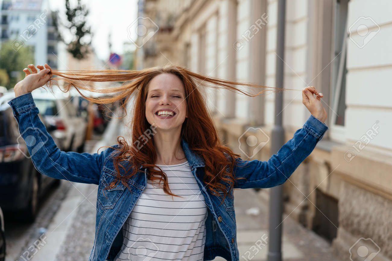 Happy young woman playing with her long red hair smiling happily at the camera as she pulls strands up in her hands on an urban street - 154410538 Happy young woman playing with her long red hair smiling happily at the camera as she pulls strands up in her hands on an urban street - 154410538