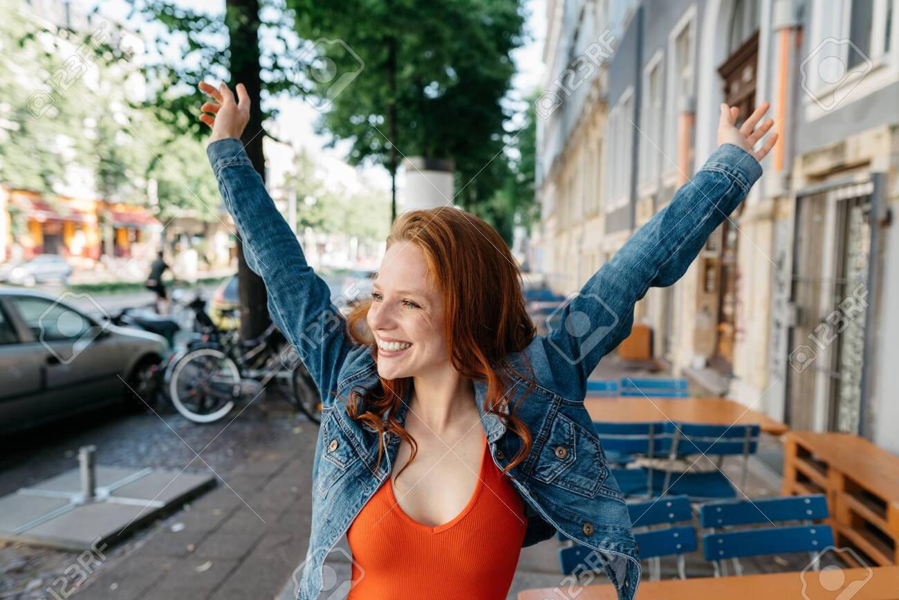 Happy casual young redhead woman rejoicing in town stretching up her arms and looking aside with a beaming smile of pleasure - 154410540 Happy casual young redhead woman rejoicing in town stretching up her arms and looking aside with a beaming smile of pleasure - 154410540