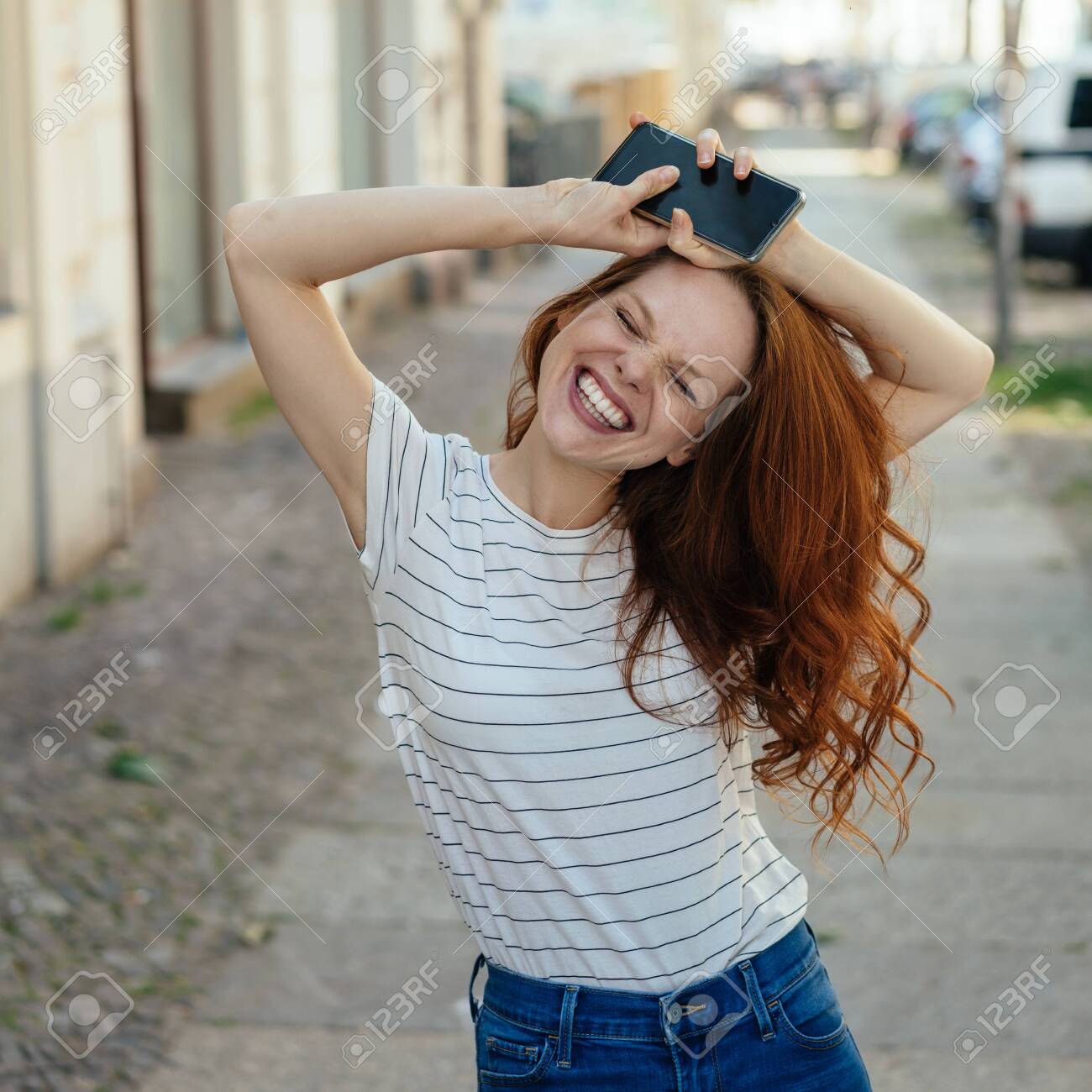 Happy young woman with beaming smile and eyes tight shut holding her mobile above her head as she grins at the camera on a quiet urban street - 154410541 Happy young woman with beaming smile and eyes tight shut holding her mobile above her head as she grins at the camera on a quiet urban street - 154410541