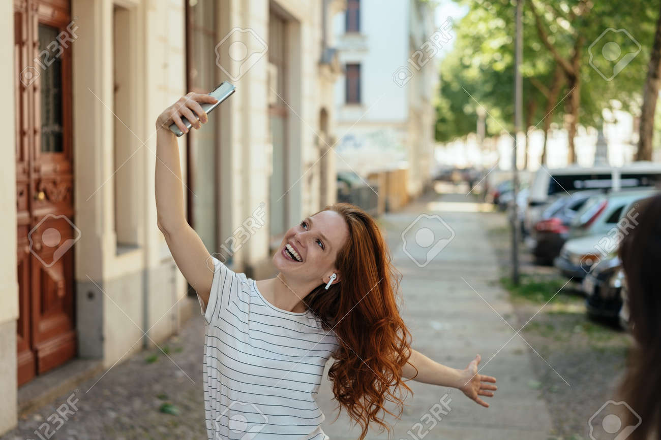 Vivacious young woman posing for a selfie on her smartphone on a quiet urban street grinning for the camera - 154410544 Vivacious young woman posing for a selfie on her smartphone on a quiet urban street grinning for the camera - 154410544