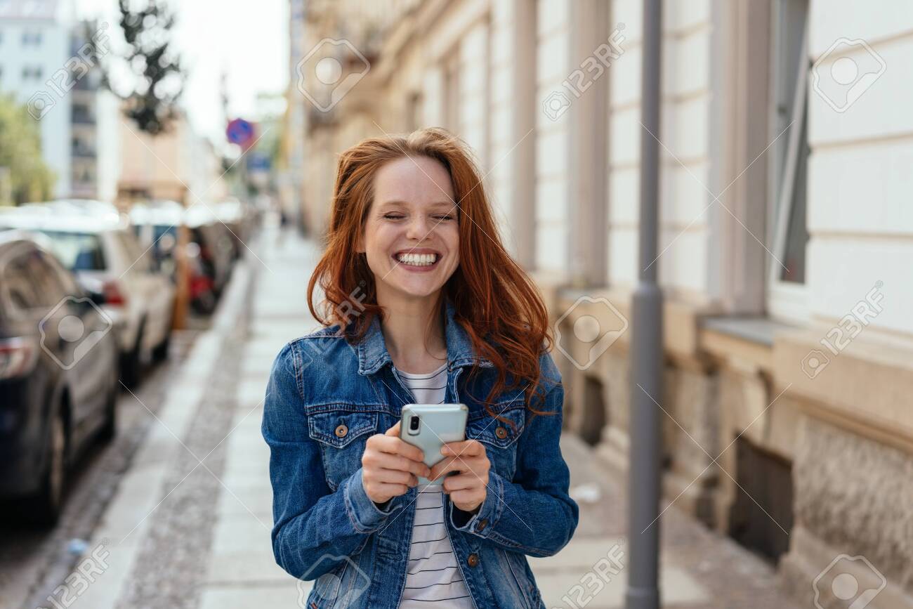 Young woman with a happy toothy grin holding a mobile in her hands on an urban street facing the camera - 154410546 Young woman with a happy toothy grin holding a mobile in her hands on an urban street facing the camera - 154410546