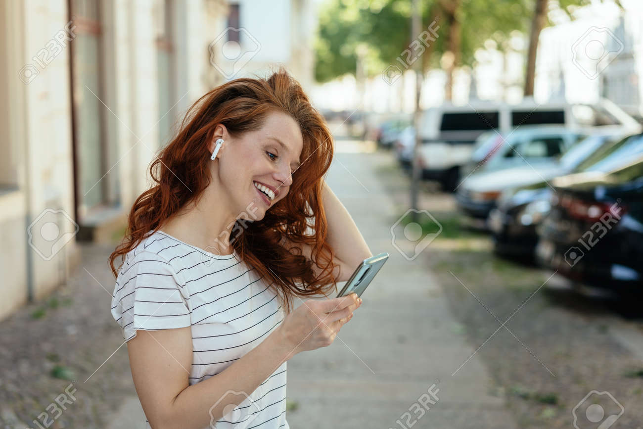 Smiling young woman listening to music or taking a call on an earbud as she looks down at her mobile phone on a quiet urban street - 154410550 Smiling young woman listening to music or taking a call on an earbud as she looks down at her mobile phone on a quiet urban street - 154410550