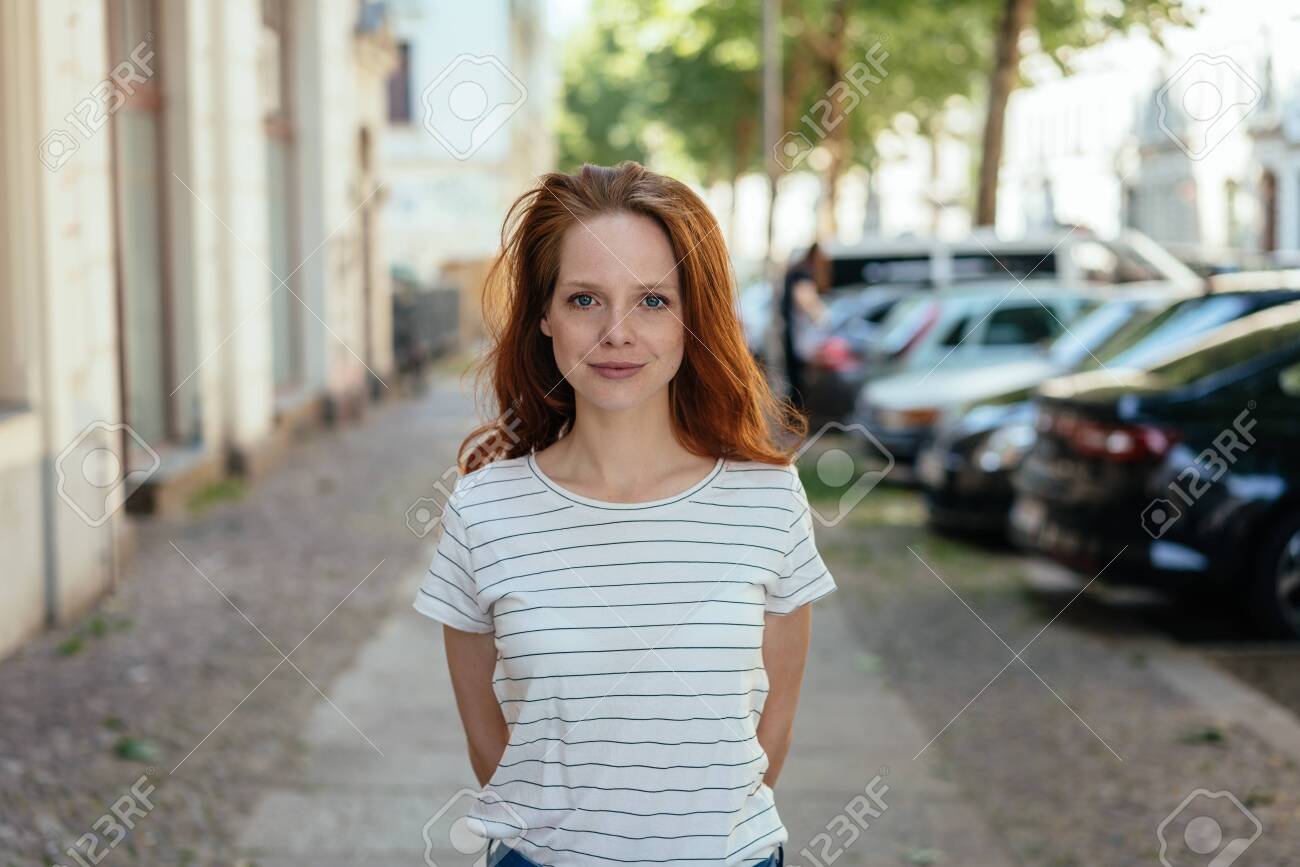 Young woman with a quiet attentive smile standing with hands behind her back on a quiet urban street in a frontal portrait - 154410575 Young woman with a quiet attentive smile standing with hands behind her back on a quiet urban street in a frontal portrait - 154410575