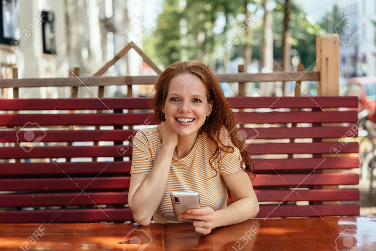 Young woman sitting at an outdoor restaurant table holding a mobile phone looking attentively at the camera - 154410576 Young woman sitting at an outdoor restaurant table holding a mobile phone looking attentively at the camera - 154410576
