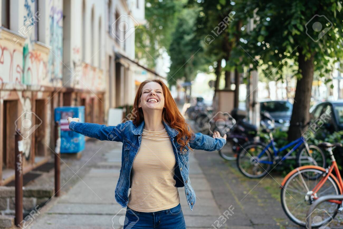 Happy young woman rejoicing with outstretched arms and a beaming smile on a sidewalk on a quiet urban street - 154410582 Happy young woman rejoicing with outstretched arms and a beaming smile on a sidewalk on a quiet urban street - 154410582