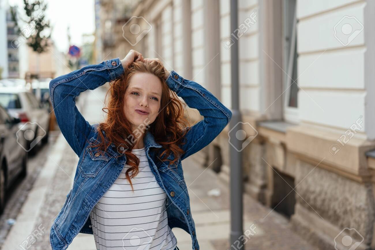 Cute charismatic young woman with playful grin standing with her hands raised on an urban street in a denim jacket - 154410583 Cute charismatic young woman with playful grin standing with her hands raised on an urban street in a denim jacket - 154410583