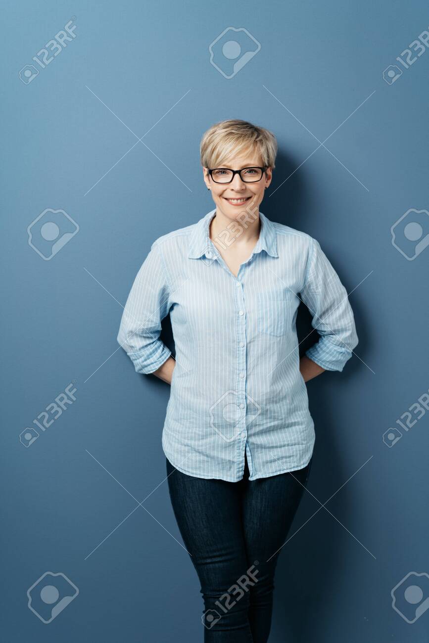 Front portrait of smiling middle-aged woman with short blond hair, wearing glasses, blue shirt and dark jeans, standing with her hands behind her back, against plain blue background in studio - 153539406 Front portrait of smiling middle-aged woman with short blond hair, wearing glasses, blue shirt and dark jeans, standing with her hands behind her back, against plain blue background in studio - 153539406