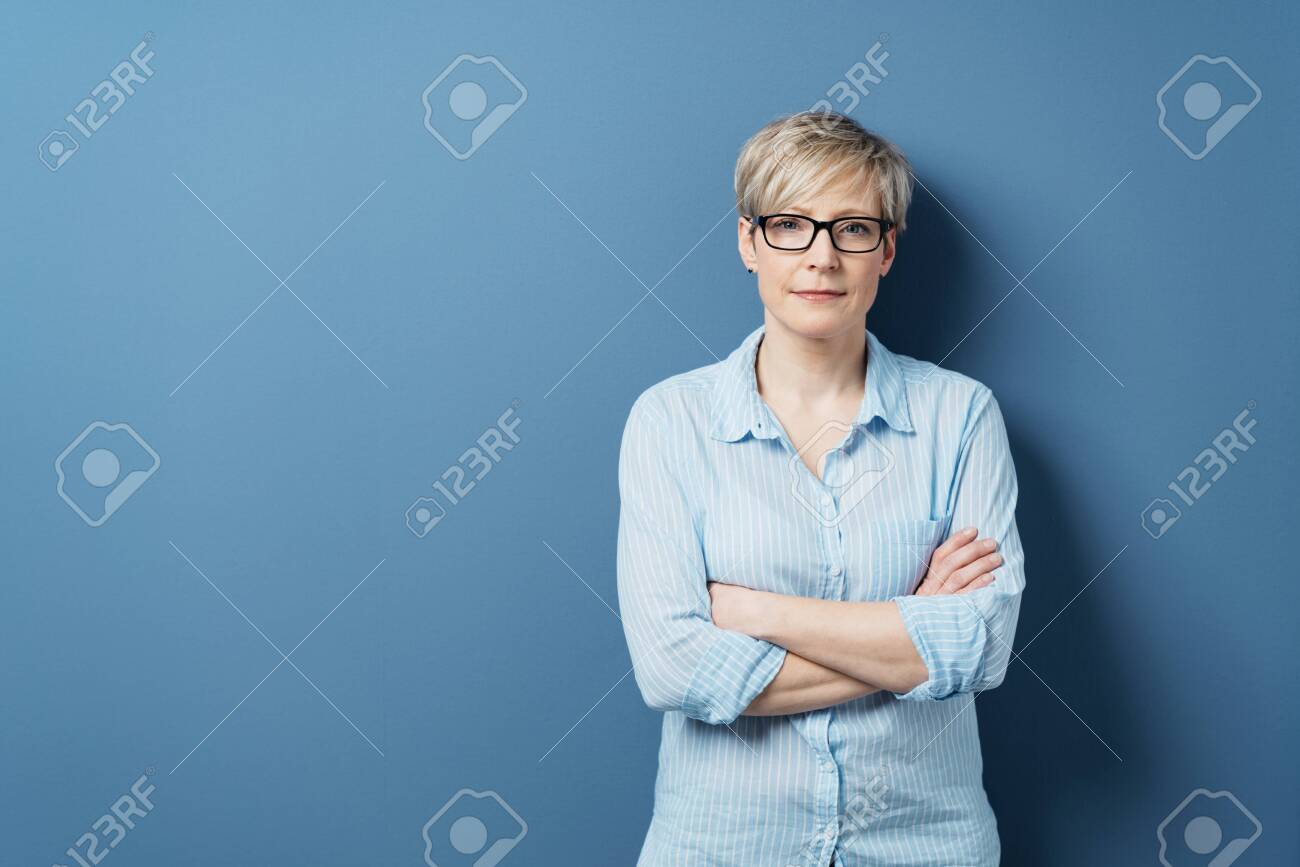 Portrait of mature blonde woman wearing glasses standing against blue background with arms crossed - 153539028 Portrait of mature blonde woman wearing glasses standing against blue background with arms crossed - 153539028