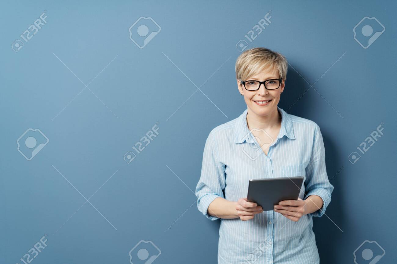 Smiling middle-aged woman with short blond hair, wearing glasses and blue shirt, standing with tablet computer in her hands and looking at camera. Front portrait on blue background with copy space - 153539368 Smiling middle-aged woman with short blond hair, wearing glasses and blue shirt, standing with tablet computer in her hands and looking at camera. Front portrait on blue background with copy space - 153539368