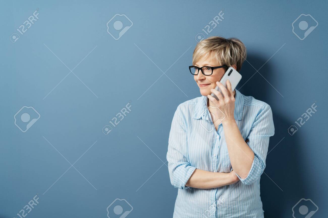 Middle-aged woman with short blond hair, in glasses and blue shirt, standing and looking aside while talking by the phone. Half-length front portrait against blue background in studio with copy space - 153539388 Middle-aged woman with short blond hair, in glasses and blue shirt, standing and looking aside while talking by the phone. Half-length front portrait against blue background in studio with copy space - 153539388