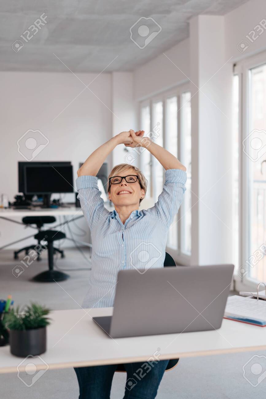 Happy businesswoman stretching her arms above her head with a beaming smile seated at a laptop in a high key office with copy space - 153539467 Happy businesswoman stretching her arms above her head with a beaming smile seated at a laptop in a high key office with copy space - 153539467