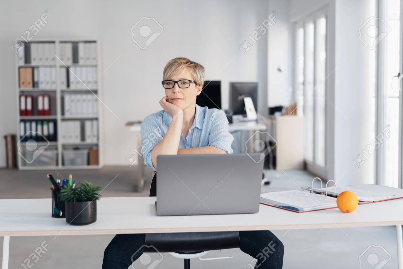 Middle-aged woman with short haircut and in glasses, sitting at laptop in empty office and thinking with sad face, propping her chin with her hand. Front portrait with copy space - 153539139 Middle-aged woman with short haircut and in glasses, sitting at laptop in empty office and thinking with sad face, propping her chin with her hand. Front portrait with copy space - 153539139