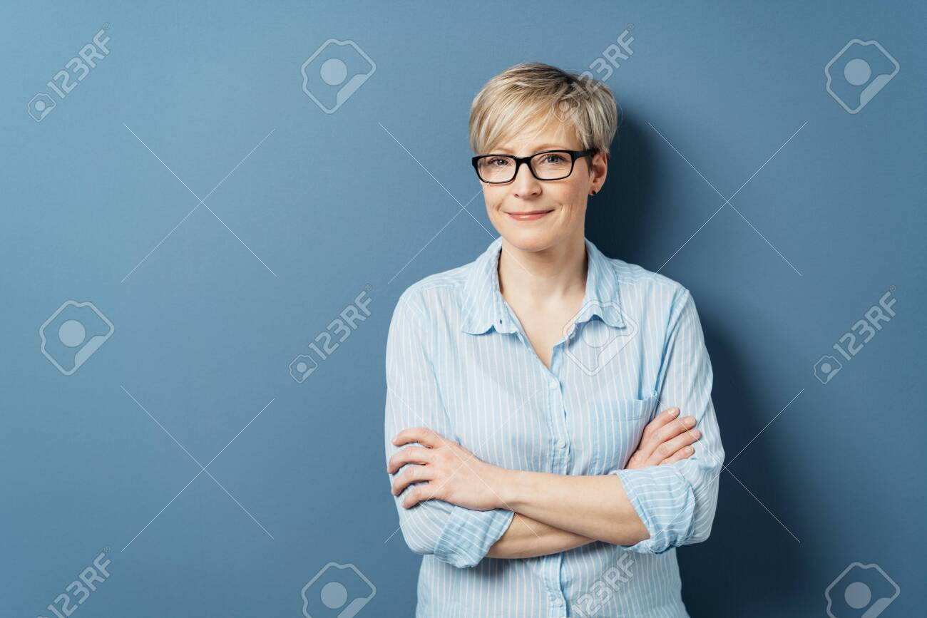 Middle-aged woman with short blond hair, in blue shirt and wearing glasses, standing with her arms crossed and looking at camera with modest smile. Half-length front portrait on blue with copy space - 153539414 Middle-aged woman with short blond hair, in blue shirt and wearing glasses, standing with her arms crossed and looking at camera with modest smile. Half-length front portrait on blue with copy space - 153539414