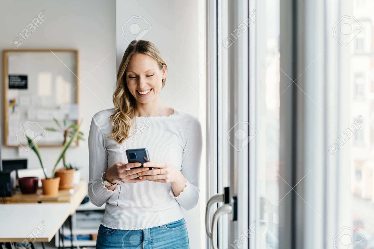 Young business woman reading a text message on her mobile with a beaming smile of pleasure as she stands in a bright office relaxing near a window with copyspace - 167565780 Young business woman reading a text message on her mobile with a beaming smile of pleasure as she stands in a bright office relaxing near a window with copyspace - 167565780