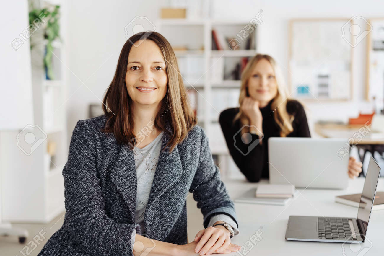 Smiling confident attentive smart middle-aged businesswoman turning to look at the camera watched by a female colleague working at a laptop in the background in a modern office - 167021653 Smiling confident attentive smart middle-aged businesswoman turning to look at the camera watched by a female colleague working at a laptop in the background in a modern office - 167021653
