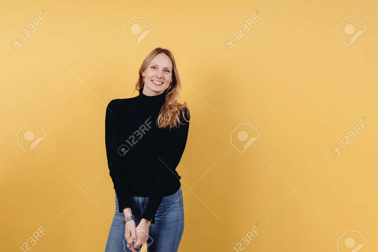 Cute vivacious young woman with long blond hair wearing jeans and a black turtle neck posing over a colorful yellow studio background with copyspace - 167021251 Cute vivacious young woman with long blond hair wearing jeans and a black turtle neck posing over a colorful yellow studio background with copyspace - 167021251