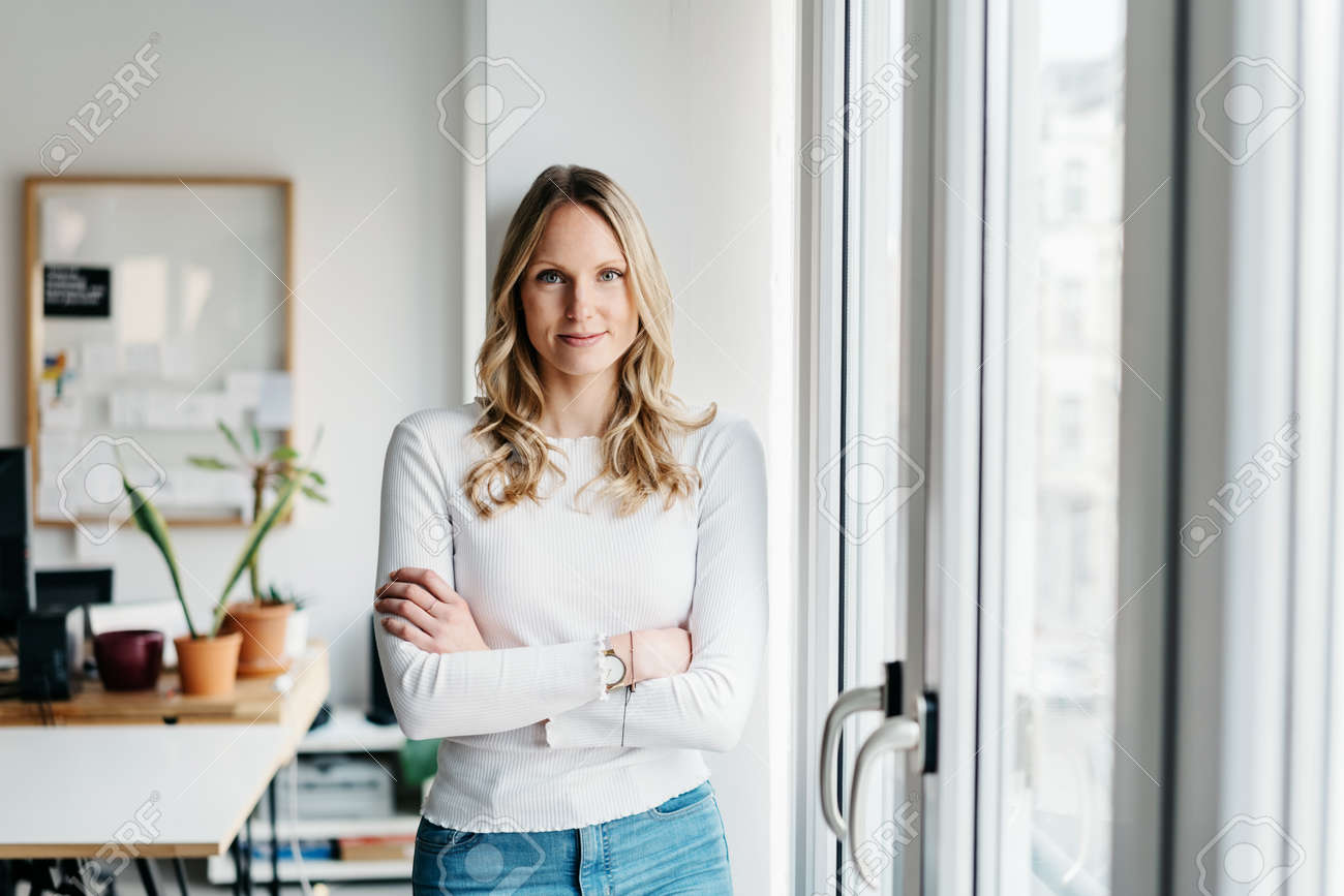 Confident friendly young woman watching the camera with folded arms as she stands alongside a bright window in a spacious office smiling happily - 167021436 Confident friendly young woman watching the camera with folded arms as she stands alongside a bright window in a spacious office smiling happily - 167021436