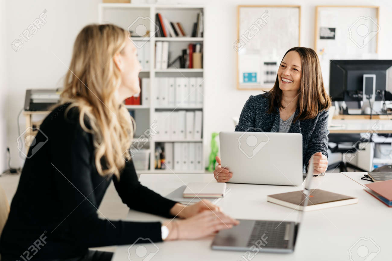 Two motivated happy businesswoman having a meeting in the office laughing and smiling as they sit at a table in a teamwork and cooperation concept - 167021410 Two motivated happy businesswoman having a meeting in the office laughing and smiling as they sit at a table in a teamwork and cooperation concept - 167021410