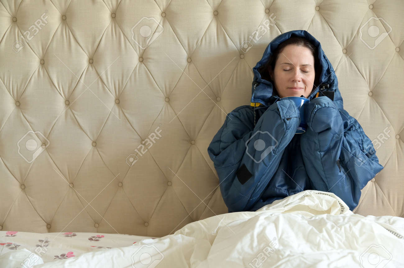 Happy adult woman wearing a very warm clothing and covered with a blanket, sitting on a double bed, having a hot drink in bedroom on a cold winter day. - 169904522 Happy adult woman wearing a very warm clothing and covered with a blanket, sitting on a double bed, having a hot drink in bedroom on a cold winter day. - 169904522